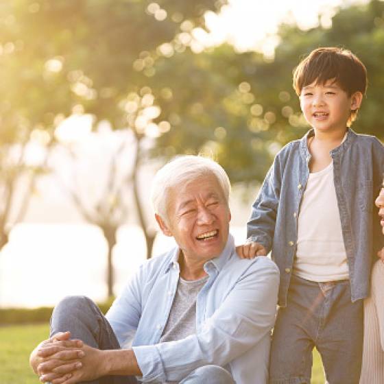 asian grandson, grandfather and grandmother sitting chatting on grass outdoors in park at dusk